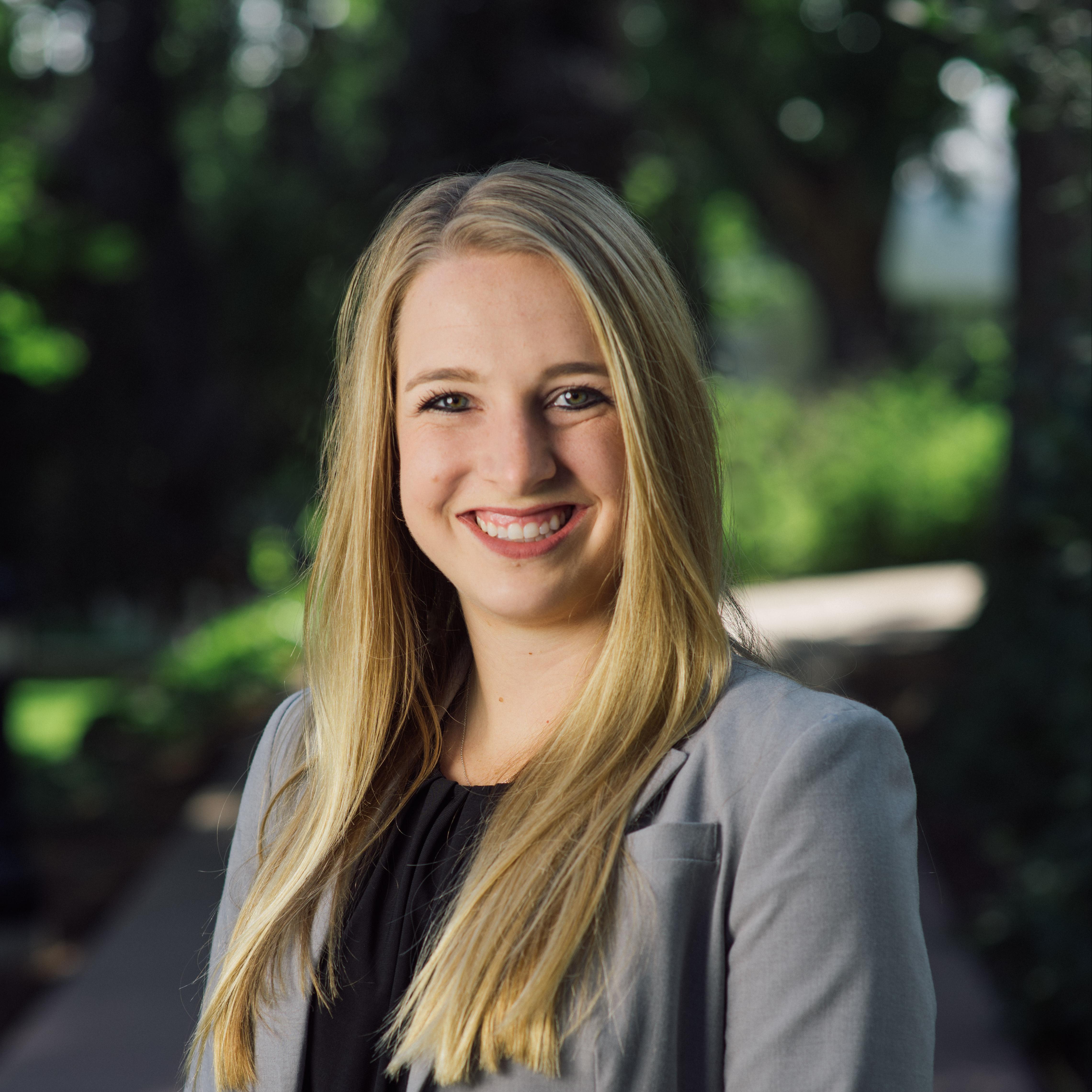 Picture of Kacie Skelton wearing a silver suit. She is smiling and has blonde hair reading her middle section of her arms. The background is of a paved trail with greenery. 
