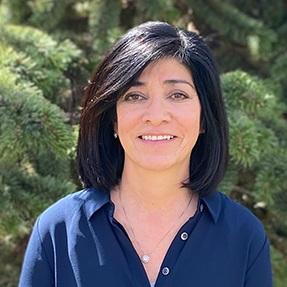 Photo of Patty Montoya. She is smiling and wearing a blue collard shirt with a necklace. There is a background of pine trees. 