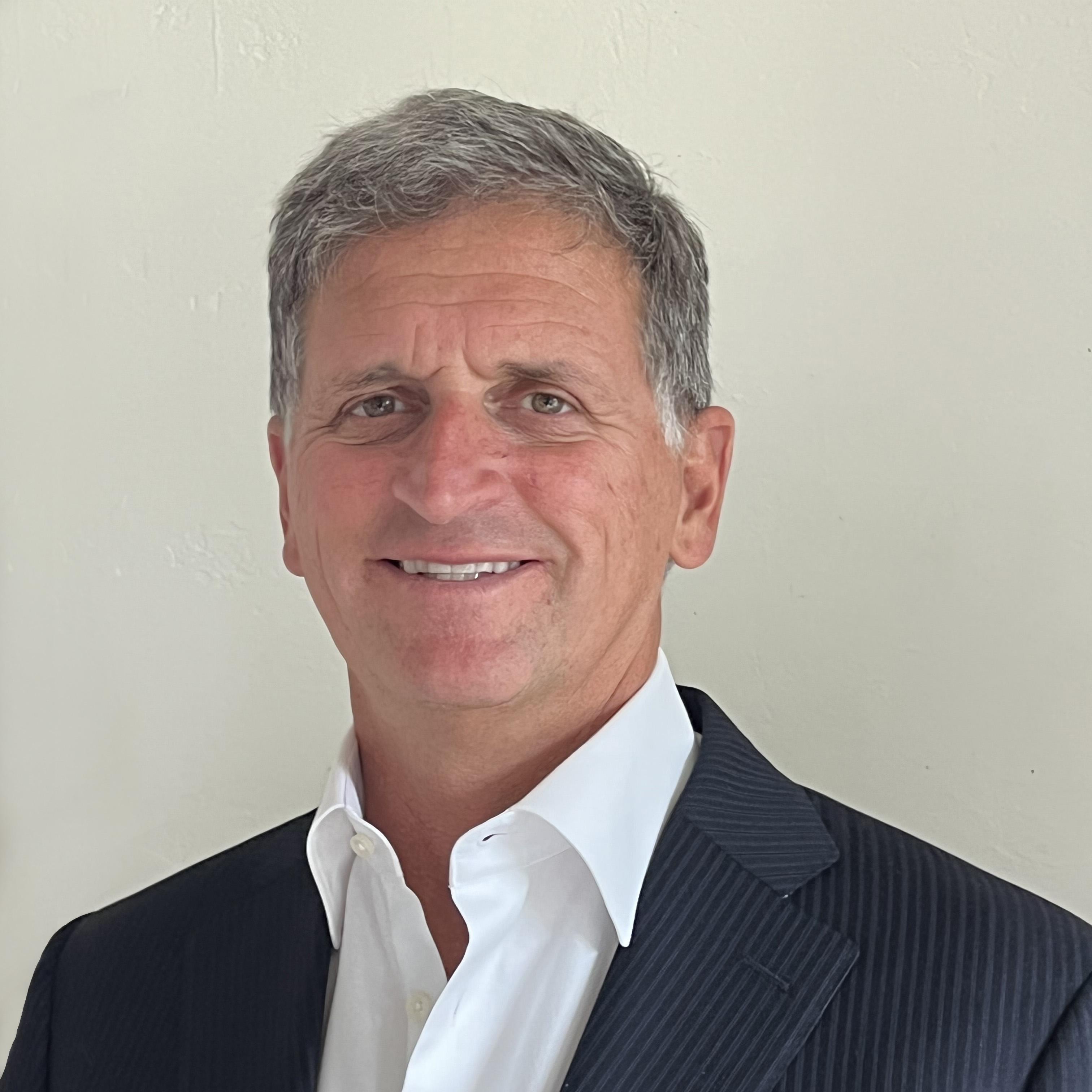 Photo of Robert Beletic smiling and wearing a black suit. He is wearing a white collar shirt and the background is a solid white-grey color. He's got white-silver hair 