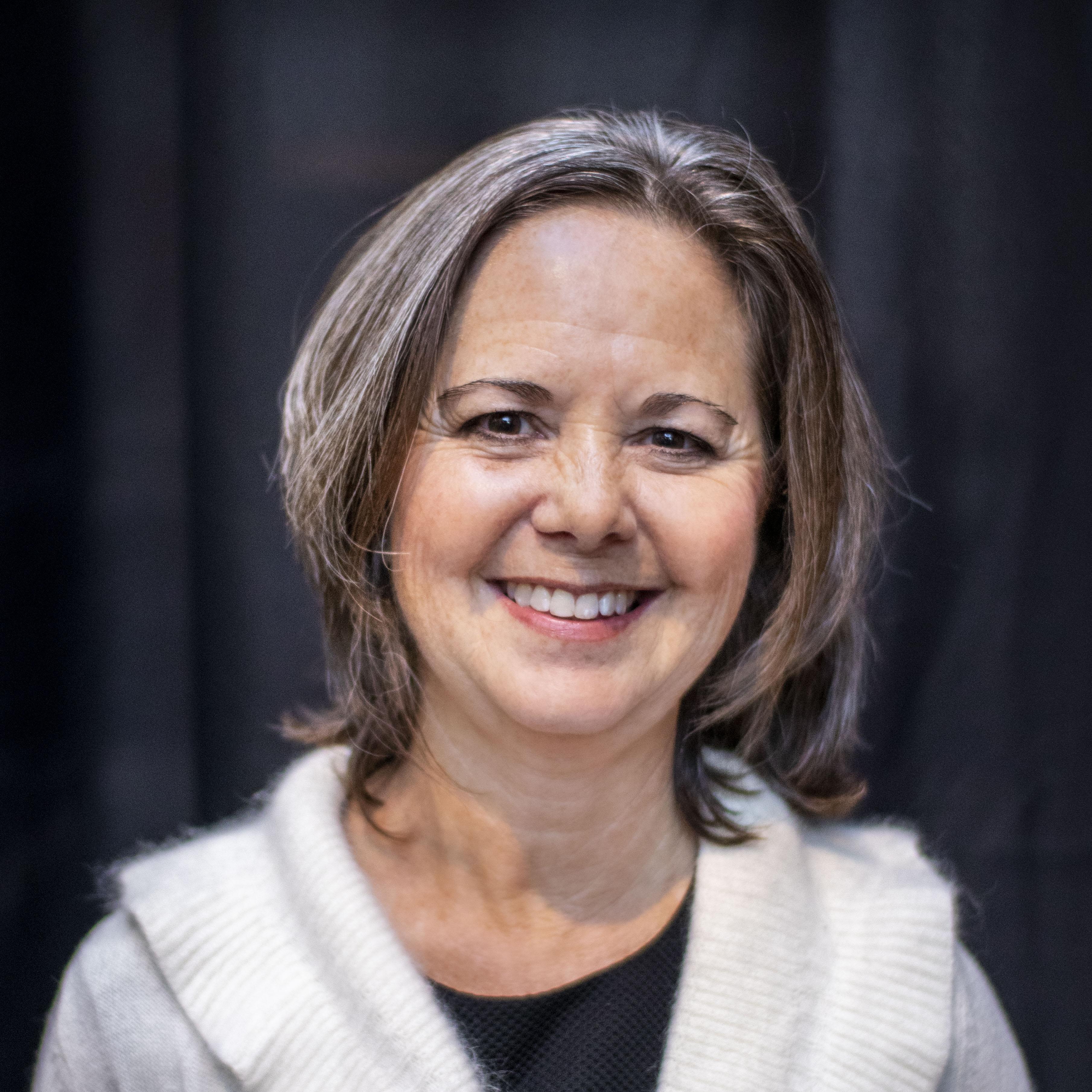 Image of Wendy from the elbows and above. She is smiling, has a white sweater, and a black shirt. Her hair is mid-length and has gray to brown hair. She has a a black curtain background 