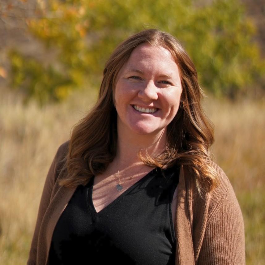 Image of Sam Taylor in nature. She has light brown hair, smiling, wearing a black shirt, has a necklace, and a light brown cardigan sweater. 