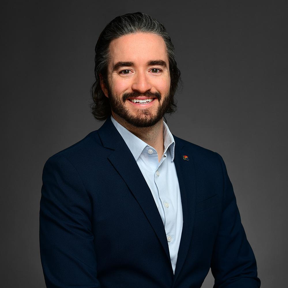 Headshot image of Christian Thomas. Christian is wearing a dark blue business suit and a light blue collard shirt. He is wearing the state of Colorado pin, has dark hair, a mustache-beard combo, and some gray hair. 