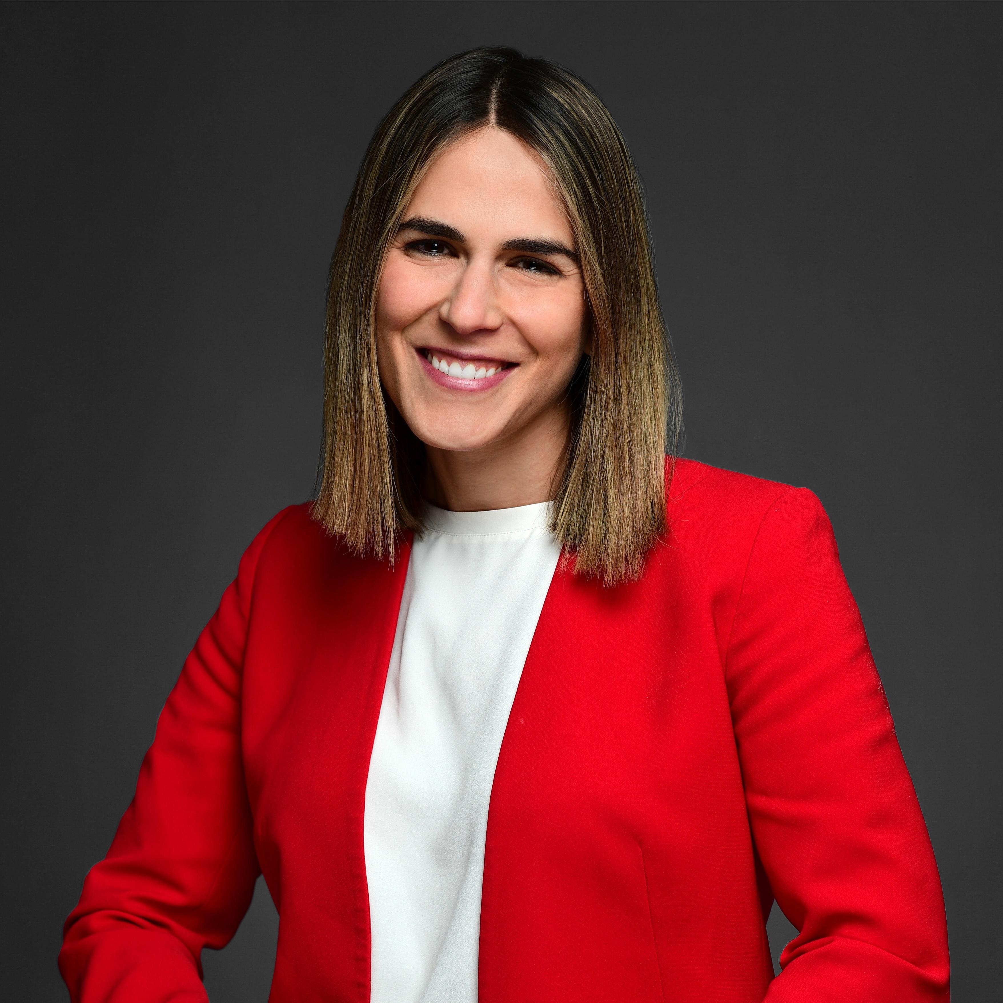 Headshot image of Eve Lieberman. She is wearing a red blazer and white shirt. She has a medium-length hair style with blonde highlights and is smiling at the camera. 