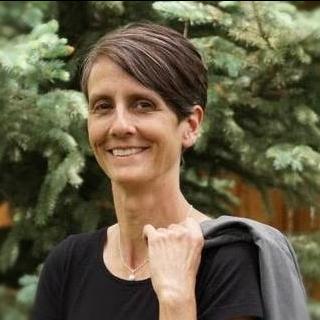 Wendy Hawthorne is smiling and looking at the camera. She has a black shirt, holding a gray jacket over her shoulder, short dark brown hair, white necklace,  and standing in front of a pine tree. 