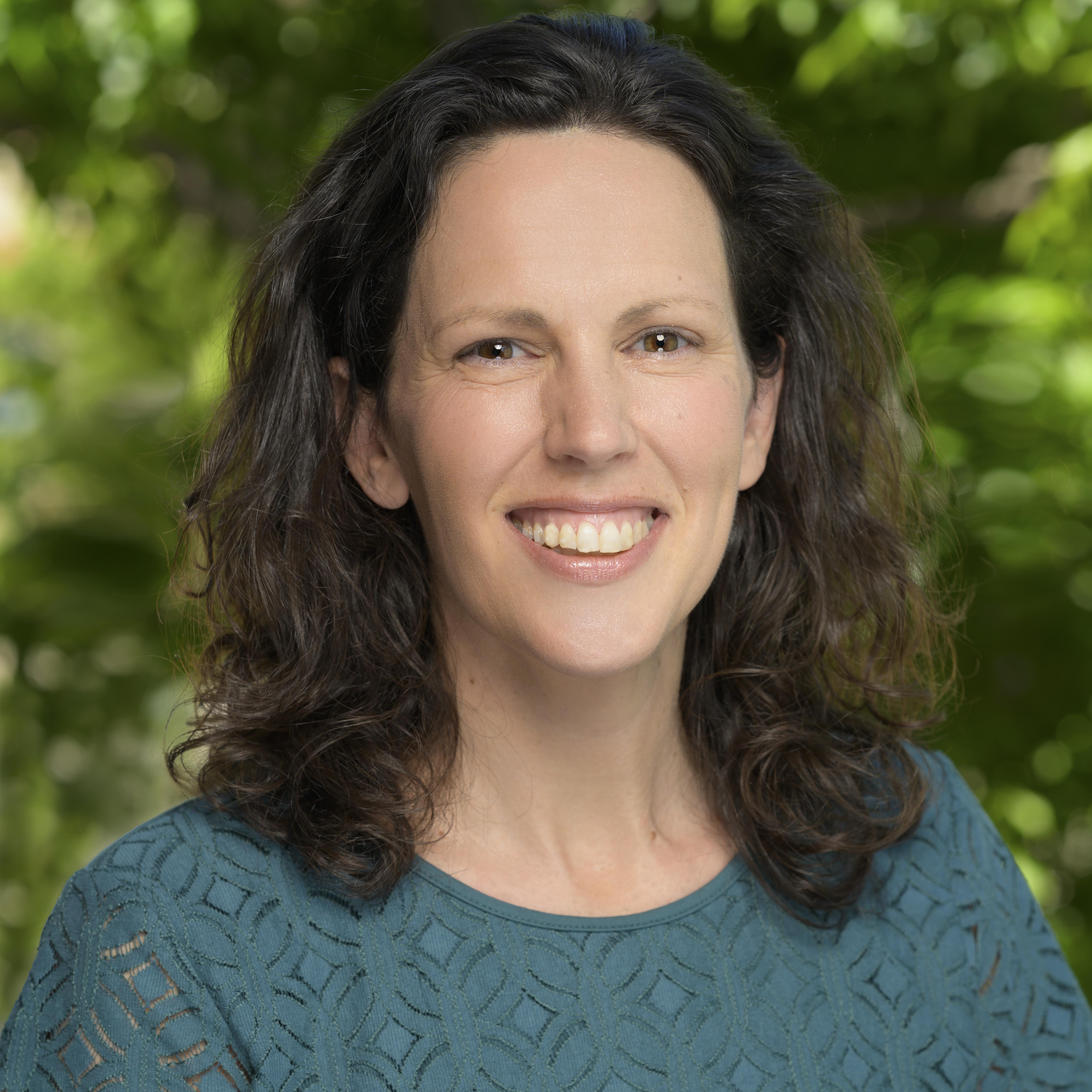 Hannah is smiling and looking at the camera. She has a geometric pattern green-blue blouse. Hannah has medium length wavey brown hair and light brown eyes. The image background is blurred green nature. 