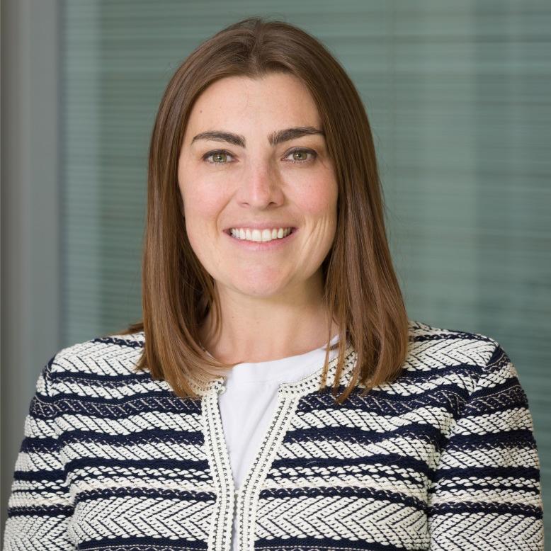 Laura has light brown hair that goes over her shoulder. She is smiling and looking at the camera. She is wearing a stripped white and blow sweater with arrow patterns. The background of the image is a window with curtains. 
