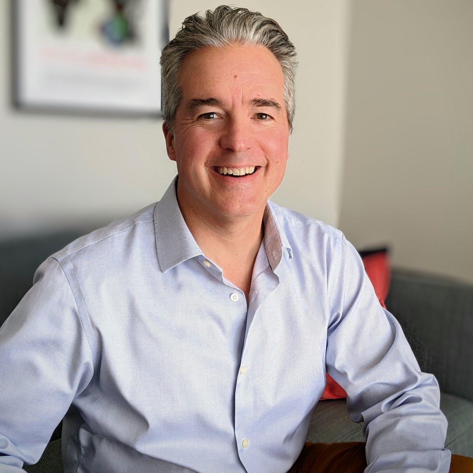 Bill is looking at the camera. He has a white collard shirt and gray white hair. He is smiling and is infront of a living room. The background has a blurred out sectional couch, pillow, and frame.  