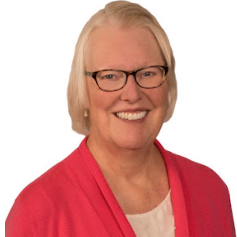 Laurie is smiling and looking at the camera directly. She has short medium light blonde hair and is wearing brown glasses with hues of yellow in them. She has small dangling earrings and a pink cardigan. She has a white shirt underneath the cardigan. 