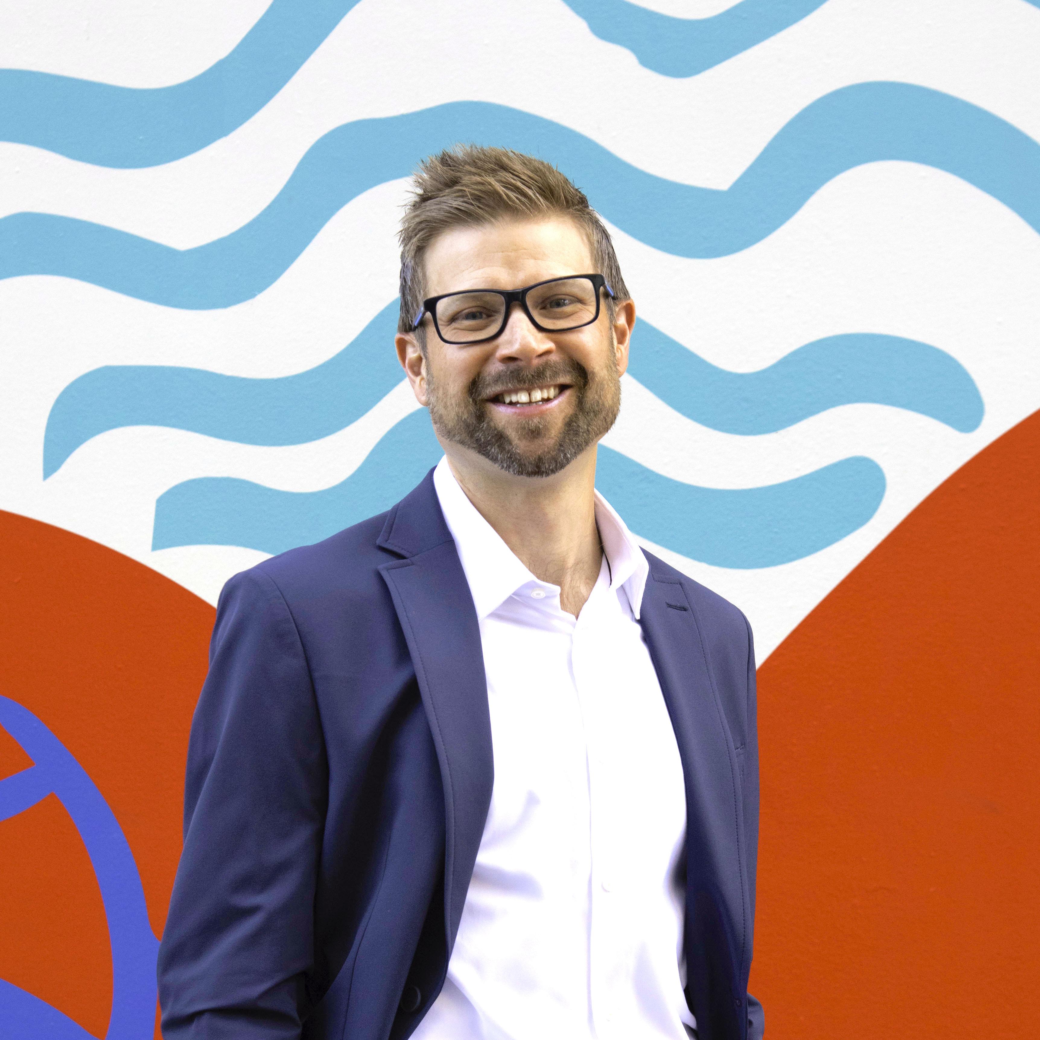 A white middle aged man with white shirt and blue suit jacket and glasses smiles in front of a red, white, and blue modern abstract mural