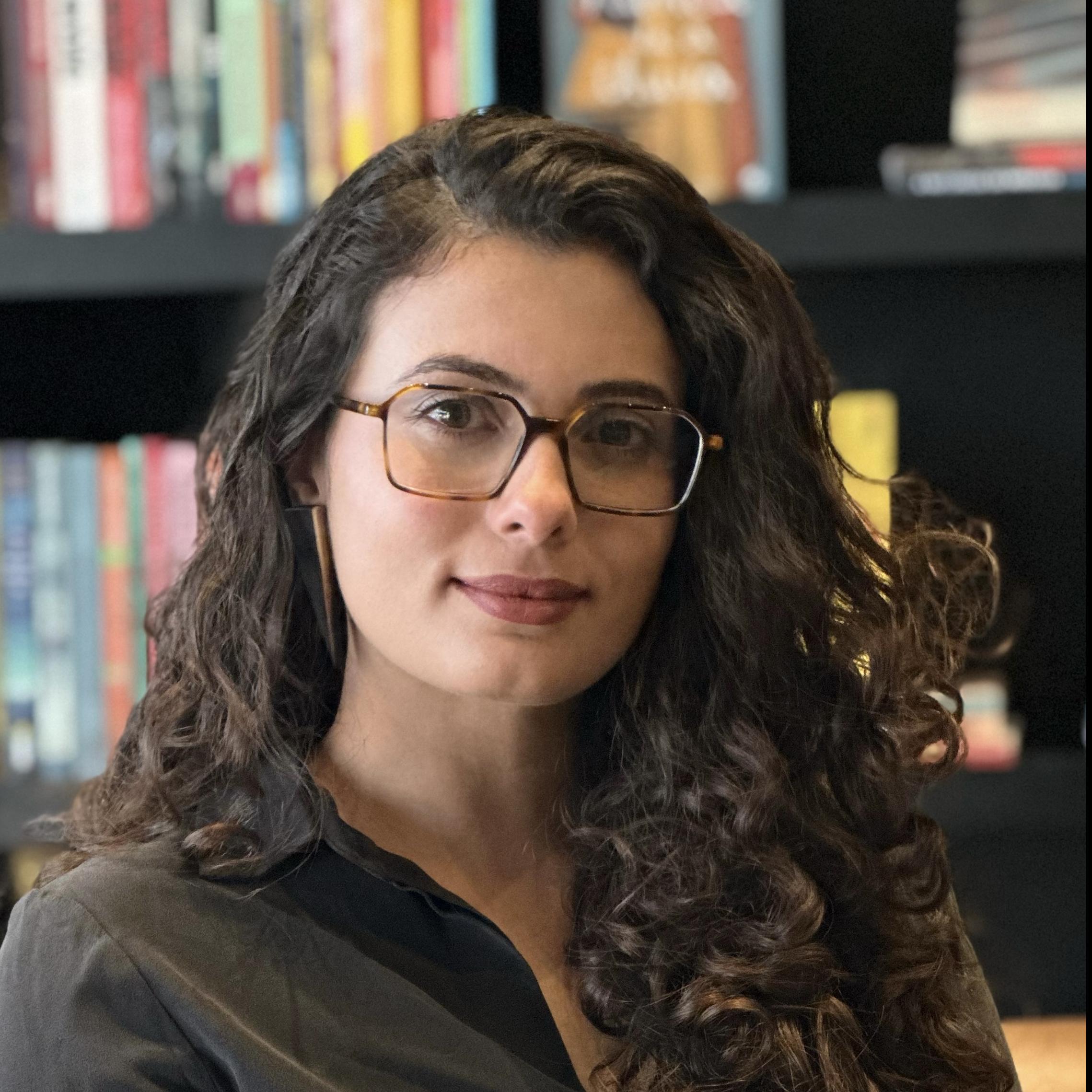 Júlia Martins Rodrigues wearing glasses and has curly hair while sitting in front of a bookshelf.