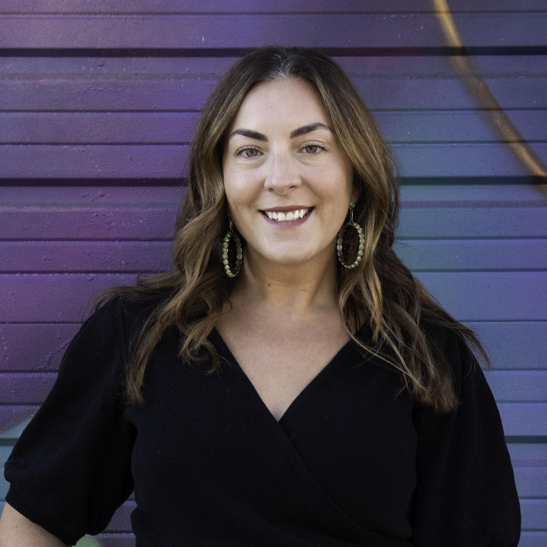 Christy Costello is standing in front of a colorful purple mural. She has curled auburn hair. She is wearing a black shirt and blue jeans.