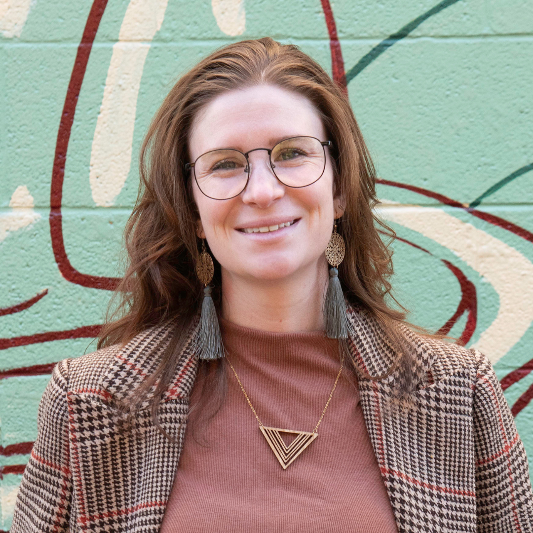 Katy Zeigler is standing in front of a light blue mural, she has auburn hair. She is wearing fall color shirt and blazer.