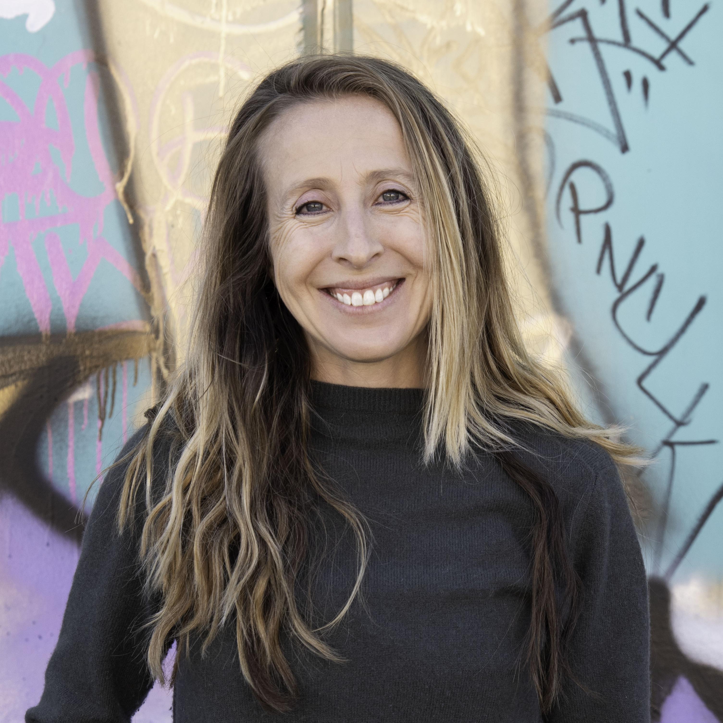 Ruth Wilson standing in front of a colorful mural, she as long brown hair with blonde highlights. She is wearing a grey long sleeve turtle neck.