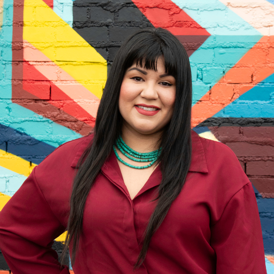 Victoria Gonzalez is standing in front of a colorful mural. She has long black hair with bangs. She is wearing a maroon jumpsuit with a turquoise necklace.