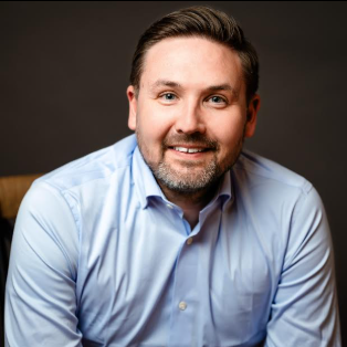 Headshot of Eric Guttormson, who is smiling, sitting on a chair with a blue shirt.