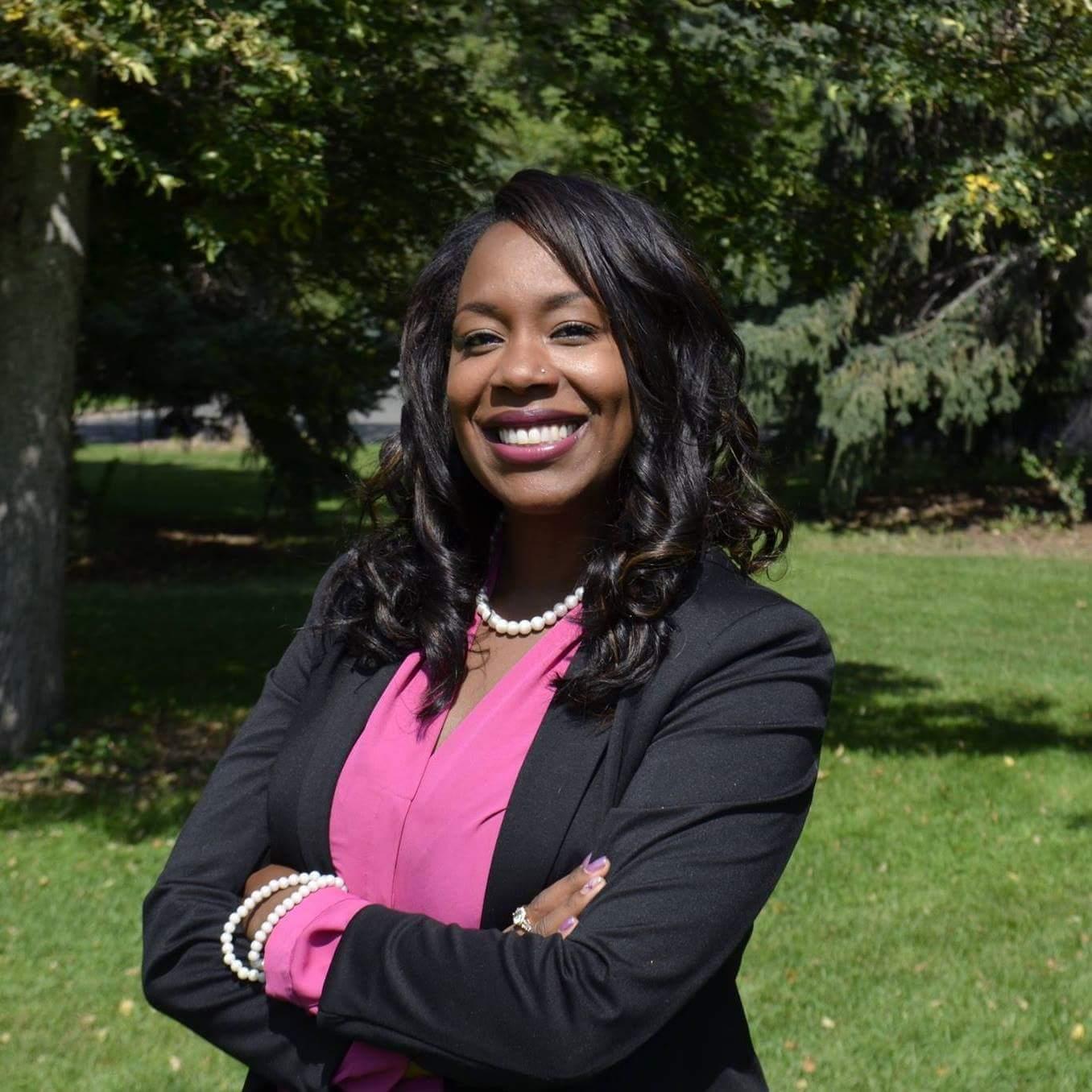 Portrait of a Jessica  smiling, wearing a black blazer and pink shirt, standing outdoors with trees and grass in the background.