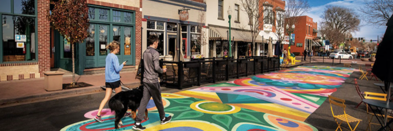 Two people walking a dark brown dog on the streets of downtown arvada, which are painted in bright colors with chalk. There is a few shops they are walking by and blue skies ahead