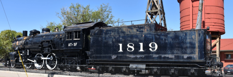 A vintage black train sits on display on tracks in front of a red silo, on a clear blue day