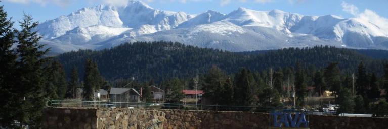 Panorama view of the City of Woodland Park with snow-capped mountains in the background, evergreen trees in the front and clear blue skies with some white fluffy clouds