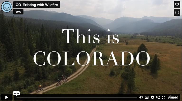 The words "This is Colorado" over a mountain valley with smoke covered peaks in the background, overlooking a trail with 3 bikers and trees and plains surrounding them