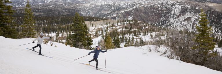 Two cross countries skiers crossing a snowy landscape with mountains in the background