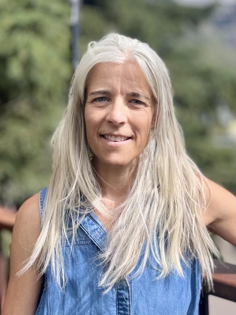 OEDIT Director of Innovative Funding for Housing Programs Hilary Cooper smiling, wearing a light denim tank top, with long grey hair in front of her shoulders.