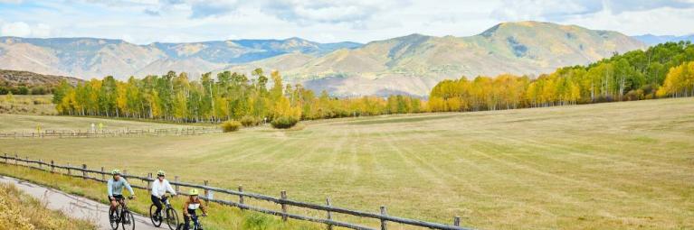 A family of four rides their bikes down a paved path, with aspen trees in the distance surrounding them, and a field of plains and dead brush