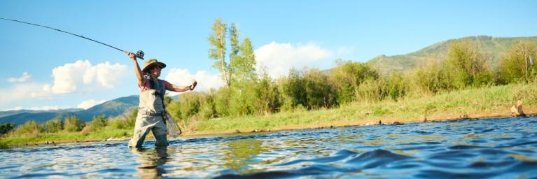 A woman fly fishing, with overalls up to her upper chest, rod back getting ready to cast forward, in blue lake water with a field of green shrubs and grass in the background
