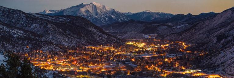 An ariel view of Glenwood Springs, with many bright lights shown coming from hotels, houses, street lights, with a snowy mountain in the background