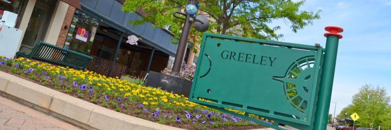 A green sign welcomes visitors to Downtown Greeley, with brown buildings in the background and a green tree