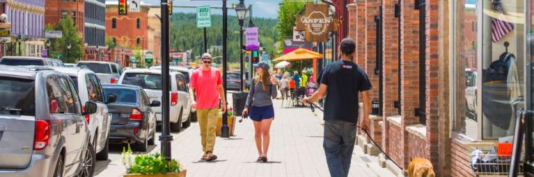 a few people walking down the street, you can see cars parked on the side of the road and shops on the right hand side