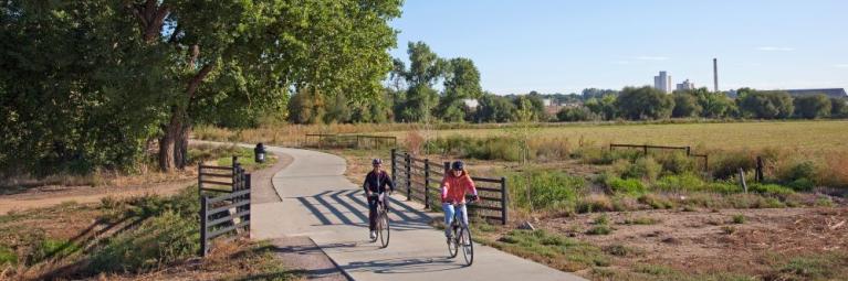 A pair of bicyclists crosses a bridge on a bike path in Longmont, with large green trees in the background, and a clear blue sky