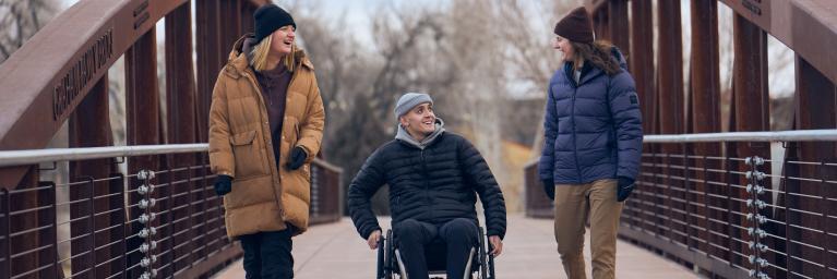 three people on a bridge wearing winter attire. Two people are walking next to a person in a wheelchair