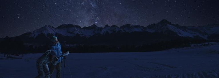 Couple looking up at the stars using a telescope with mountains in the background.