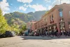 Two bicyclists ride through a downtown area during the day with mountains in the background.
