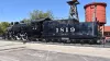 A vintage black train sits on display on tracks in front of a red silo.