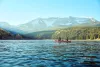 Two people canoe through a blue lake with mountains and forest in the background.
